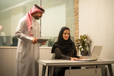 Monitoring workflow using a smart device, exchanging ideas and information, teamwork within an office, the concept of professional communication, an Arab Gulf Saudi employee standing next to the desk holding a tablet discussing with his colleague, an Arab Gulf Saudi female employee sitting in front of a laptop.