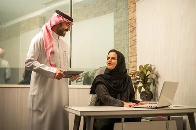 Exchanging ideas and information, monitoring workflow using smart devices, teamwork within the office, the concept of professional communication, a Saudi Gulf Arab employee standing next to the desk holding a tablet discussing with his colleague, a Saudi Gulf Arab female employee sitting in front of a laptop.
