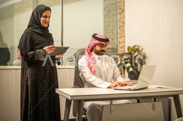 مراجعة البيانات والمعلومات، مناقشة مهام العمل اليومية, مفهوم التواصل المهني في بيئة العمل, موظفة عربية خليجية سعودية ترتدي العباءة و الحجاب تمسك بيديها جهاز التابلت تقف بجانب زميلها، استخدام الحاسب المحمول لانجاز العمل