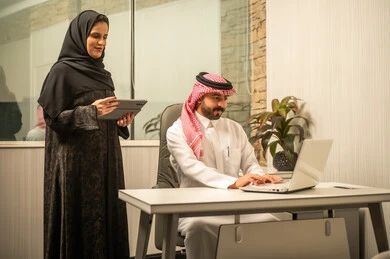 مراجعة البيانات والمعلومات، مناقشة مهام العمل اليومية, مفهوم التواصل المهني في بيئة العمل, موظفة عربية خليجية سعودية ترتدي العباءة و الحجاب تمسك بيديها جهاز التابلت تقف بجانب زميلها، استخدام الحاسب المحمول لانجاز العمل