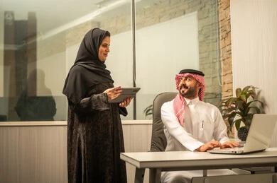 Discussion of daily work tasks, the concept of professional communication in the workplace, reviewing data and information, an Arab Gulf Saudi female employee wearing an abaya and hijab holding a tablet in her hands talking to her colleague, using a laptop to complete tasks.