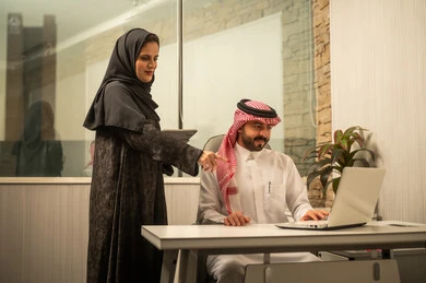 مفهوم التعاون والتواصل في بيئة العمل, استخدام التقنية في إنجاز المهام الإدارية, رجل عربي خليجي سعودي يرتدي الثوب الابيض و الشماغ  يجلس خلف المكتب ويستخدم الحاسب المحمول برفقة زميلتة, الاشارة الى شاشة الحاسب أثناء مناقشة العمل