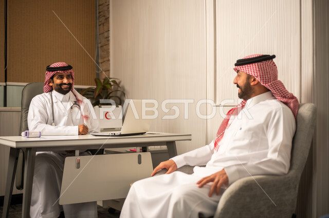 تقديم الخدمات الصحية للمرضى، مفهوم الاستشارة الطبية في المركز الطبي، طبيب عربي خليجي سعودي يرتدي الثوب الابيض و الشماغ و المعطف الطبي يجلس خلف المكتب ويستمع للمريض