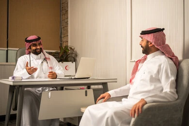 مهن ووظائف طبيه، مفهوم الاستشارة الطبية في المركز الطبي، طبيب عربي خليجي سعودي يرتدي الثوب الابيض و الشماغ و المعطف الطبي يجلس خلف المكتب ويستمع للمريض