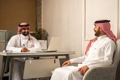 مفهوم الاستشارة الطبية، الرعاية الصحية الحديثة, متابعة الحالة الصحية للمريض, طبيب عربي خليجي سعودي يرتدي الثوب و الشماغ و المعطف الطبي  يقوم بتدوين ملاحظات المريض