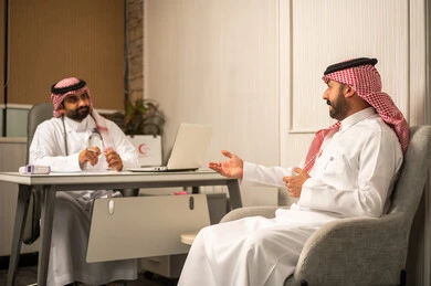 العمل في القطاع الصحي، مفهوم الاستشارة الطبية في المركز الطبي، طبيب عربي خليجي سعودي يرتدي الثوب الابيض و الشماغ و المعطف الطبي يجلس خلف المكتب ويستمع للمريض