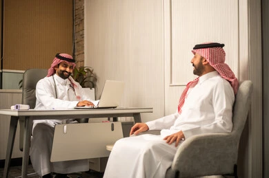 العمل في المراكز والمستشفيات، دراسة و مراجعة حالات المرضى، الخدمات الطبية الحديثة, استشارة طبية داخل عيادة, طبيب عربي خليجي سعودي يستخدم الحاسب المحمول أثناء الحديث مع المريض