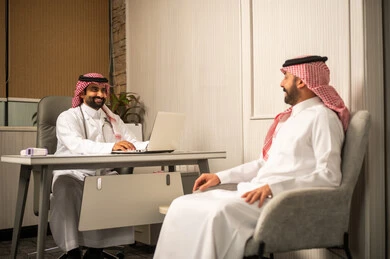 Working in the healthcare sector, medical consultation inside the clinic, a Saudi Gulf Arab doctor wearing a white thobe, a ghutrah, and a lab coat sits behind the desk and uses a laptop while talking to the patient.