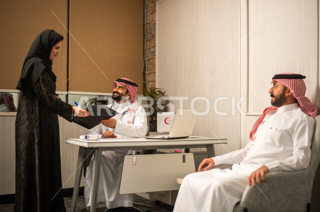 Medical consultation inside the clinic, the concept of medicine and healthcare, following up on medical and administrative procedures, a veiled Saudi Gulf Arab nurse wearing an abaya handing over medical documents to the doctor.