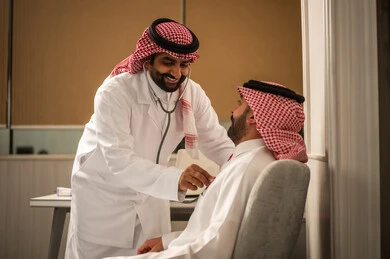 طبيب عربي خليجي سعودي يرتدي الثوب الابيض و الشماغ و المعطف الطبي يقوم باستخدام سماعه الفحص الطبية للمريض، المتابعة الدورية لفحص نبضات القلب، مفهوم الطب والرعاية الصحية، قياس معدل ضربات القلب