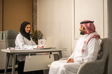 مفهوم التواصل بين الطبيب والمريض، الرعاية الصحية الحديثة, جلسة استشارة طبية داخل عيادة, طبيبة عربية خليجية سعودية محجبة ترتدي المعطف الابيض تستخدم الحاسب المحمول، مناقشة الحالة الصحية مع المريض