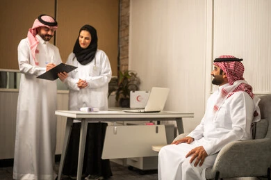 استشارة طبية داخل العيادة، طبيب وطبيبة عربيان خليجيان سعوديان يرتديان الزي الطبي يناقشان ملف المريض، مريض عربي خليجي سعودي يرتدي الثوب الابيض و الشماغ يجلس على كرسي الفحص، مفهوم الرعاية الصحية ، كتابة و تدوين الملاحظات