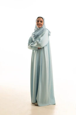 النظر إلى الكاميرا بإيماءات السرور والثقة بالنفس، مفهوم الاناقه و الاهتمام بالمظهر الخارجي، بورتريه لإمرأة عربية خليجية سعودية ترتدي الحجاب والعباءة تقف بأيدي متقاطعه، صورة الجسم بالطول الكامل، خلفية بيضاء