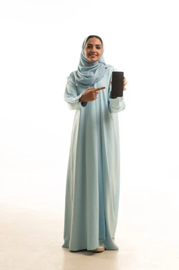 استخدام الأجهزة التقنية الحديثة للتسلية والترفيه, النظر إلى الكاميرا بإيماءات السعادة و السرور, بورتريه لشابة عربية خليجية عمانيه مبتسمة محجبة ترتدي العباءة تمسك بيدها الهاتف المحمول, عرض شاشة سوداء فارغة عبر الجوال و تشير اليها, الجسم بالطول الكامل، خلفية بيضاء