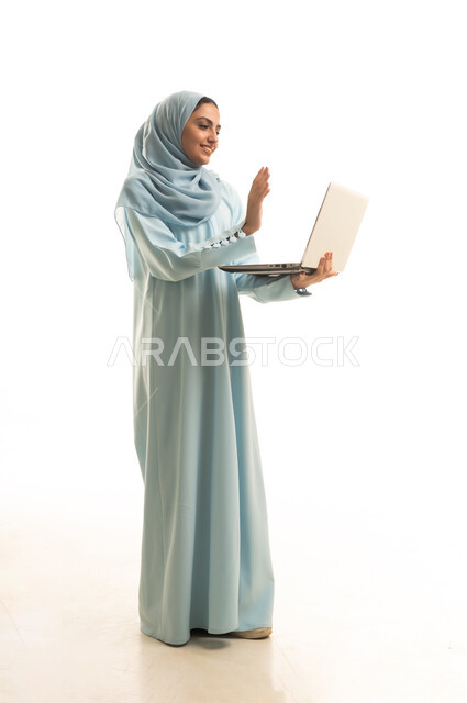 اجراء مكالمة فيديو،رفع كف اليد بتعابير التحية و السلام، ادارة العمل عن بعد، استخدام جهاز تقني حديث ومتطور، بورتريه لامرأة عربية خليجية سعودية محجبة ترتدي العباءة تعمل على جهاز الحاسوب المحمول, الجسم بالطول الكامل، خلفية بيضاء
