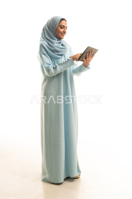 تعابير وايماءات الاندماج، استخدام التقنية الحديثه المتطورة في الحياة العملية عبر الجهاز اللوحي، بورتريه لشابة عربية خليجية سعودية ترتدي الحجاب والعباءة تقوم بالعمل على جهازالتابلت، صورة الجسم بالطول الكامل، خلفية بيضاء
