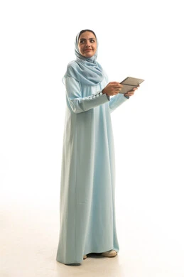 النظر الى شيء ما، استخدام التقنية الحديثه المتطورة في الحياة العملية عبر الجهاز اللوحي، بورتريه لشابة عربية خليجية سعودية ترتدي الحجاب والعباءة تقوم بالعمل على جهازالتابلت، صورة الجسم بالطول الكامل، خلفية بيضاء