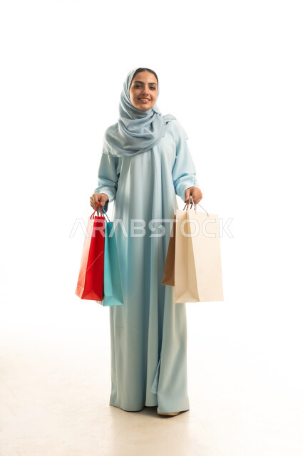 النظر بإيماءات الفرح و السرور ، الحصول على العروض و الحسومات، التسوق و شراء المستلزمات و الهدايا , بورتريه لامرأة عربية خليجية سعودية ترتدي العباءة و الحجاب تحمل أكياس المشتريات بيديها, صورة للجسم بالطول الكامل , خلفية بيضاء