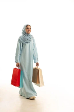 الحصول على العروض و الحسومات، التسوق و شراء المستلزمات و الهدايا , بورتريه لامرأة عربية خليجية سعودية ترتدي العباءة و الحجاب تحمل أكياس المشتريات بيديها , النظر بإيماءات الفرح و السرور , صورة للجسم بالطول الكامل , خلفية بيضاء