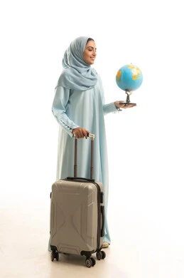 مفهوم السياحة في إجازة الصيف، الفرح لقضاء رحلة صيفية ممتعة، بورتريه لامرأة عربية سعودية خليجية ترتدي العباءة و الحجاب تقف باستقامة و تقوم بجر حقيبة الامتعة و تحمل بيدها مجسم الكرة الارضية, ايماءات السعادة والسرور، خلفية بيضاء