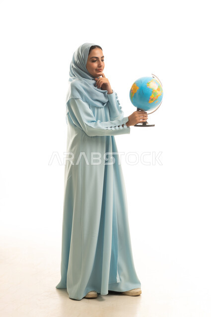 Identifying the location of Arab countries on the world map, research and exploration about the universe, a portrait of a Saudi Arabian Gulf woman wearing an abaya and hijab holding a globe in her hands, the concept of trips and tourist tours, full-body, white background.