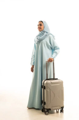 الاستمتاع بالرحلات السياحية في وقت العطلة، التجهيز للسفرات الممتعة, بورتريه لامرأة عربية خليجية سعودية محجبة ترتدي العباءة تمسك بيدها حقيبة سفر ، النظر بإيماءات السعادة والسرور، الجسم بالطول الكامل, خلفية بيضاء