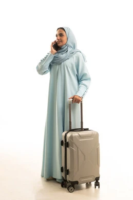 Communication with family and friends, a portrait of a Gulf Arab Saudi woman wearing an abaya holding a travel bag and a mobile phone making a phone call, using devices, technologies, and travel applications, full-body image, white background.