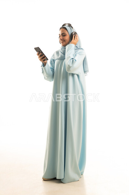 Listening to songs and music, using a modern technical device, a portrait of a Saudi Gulf Arab woman wearing a hijab and abaya, wearing headphones and holding a mobile phone, expressions of joy and happiness, full-body, white background.