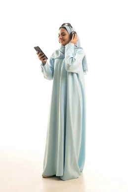 الاستماع الى الاغاني والموسيقى، استخدام جهاز تقني حديث، بورتريه لإمرأة عربية خليجية سعودية ترتدي الحجاب والعباءة  تضع سماعات الهيدفون تمسك بيدها الهاتف المحمول، تعابير السرور والسعادة، الجسم بالطول الكامل، خلفية بيضاء