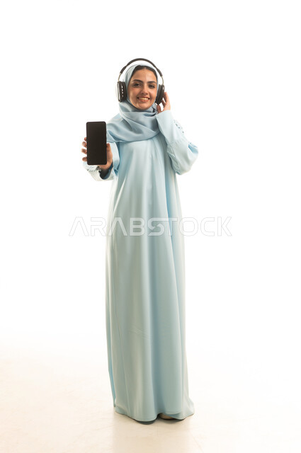 Looking at the camera and viewing a blank black screen via mobile phone, using modern and advanced technology and devices, a portrait of a smiling, veiled Saudi Arabian Gulf woman wearing an abaya and headphones, full-length body, white background