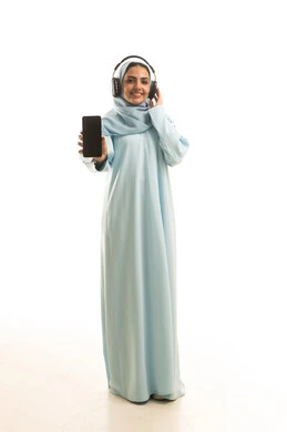 Looking at the camera and viewing a blank black screen via mobile phone, using modern and advanced technology and devices, a portrait of a smiling, veiled Saudi Arabian Gulf woman wearing an abaya and headphones, full-length body, white background