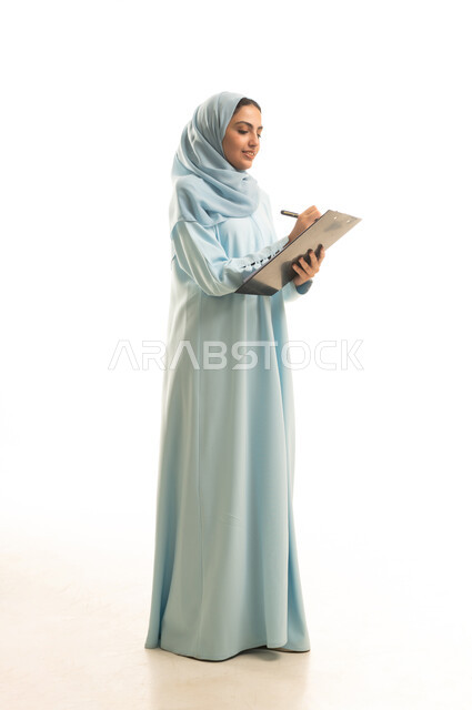 Taking notes and recording information on paper, organizing and managing business, portrait of a veiled Saudi Arabian woman wearing an abaya, holding a pen and writing notes, writing down daily tasks and goals, full-length body, white background