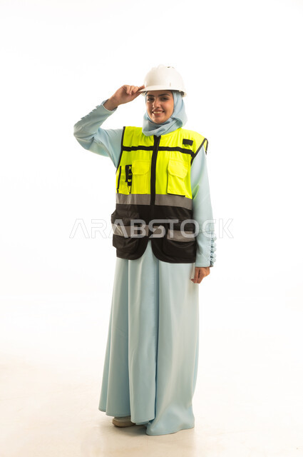 الوقوف بايماءت الثقة، مفهوم ادارة المشاريع الهندسية, وظائف ومهن نسائية, العمل في القطاع الهندسي, بورتريه لمهندسة معمارية عربية خليجية سعودية ترتدي خوذة الحماية والسترة تنظر الى الكاميرا بتعابير السرور، الجسم بالطول الكامل، خلفية بيضاء