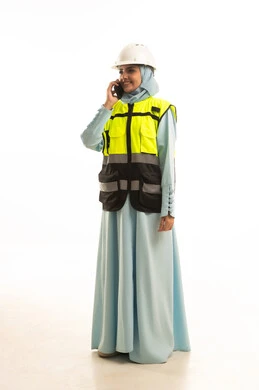 الاستشارة الهاتفية والمتابعة مع فريق العمل، دمج التكنولوجيا مع الأعمال المكتبية، بورتريه لمهندسة عربية خليجية سعودية ترتدي الخوذة البيضاء والسترة الخاصة بالعملتجري مكالمة عبرجهاز الجوال, الجسم بالطول الكامل، خلفية بيضاء