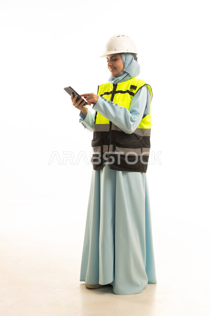 The use of modern technologies and techniques in the engineering sector, construction by the hands of the nation's sons, the concept of engineering and architectural constructions, a portrait of a Saudi Gulf Arab engineer wearing a helmet and safety vest browsing a mobile phone, full-body, white background.