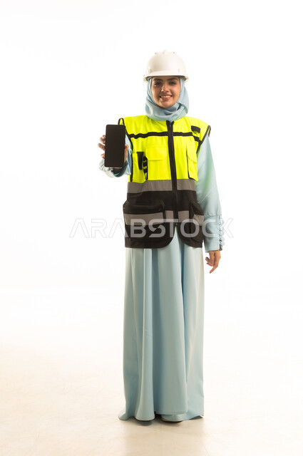 عرض شاشة سوداء فارغة عبر الهاتف المحمول, ايماءات تدل على السعادة والفرح, بورتريه لمهندسة عربية سعودية خليجية سعودية مبتسمة ترتدي العباءة و الخوذة و سترة الحماية تمسك بيديها الجوال ،مخططات وتصاميم هندسية، الجسم بالطول الكامل ، خلفية بيضاء