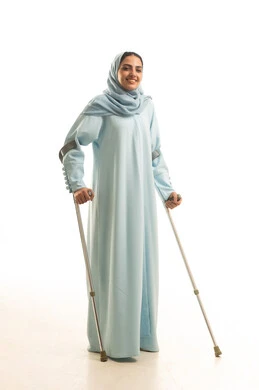 تحسين جودة حياة ذوي الاحتياجات الخاصة ودمجهم في المجتمع، استخدام العكازات الطبية للمساعدة على الحركة والتنقل، أسلوب حياة ذوي الهمم، بورتريه لامره عربيه خليجيه سعودية محجبة ترتدي العباءة تقف و تستندعلى عصا حديدية، الجسم بالطول الكامل، خلفية بيضاء