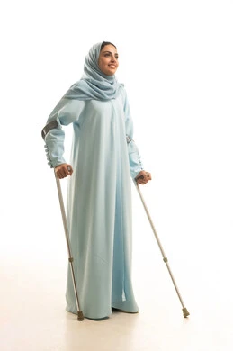استخدام العكازات الطبية للمساعدة على الحركة والتنقل، أسلوب حياة ذوي الهمم، تحسين جودة حياة ذوي الاحتياجات الخاصة ودمجهم في المجتمع، بورتريه لامره عربيه خليجيه سعودية محجبة ترتدي العباءة تقف و تستندعلى عصا حديدية، الجسم بالطول الكامل، خلفية بيضاء