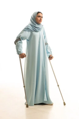 The lifestyle of people with determination, using medical crutches to assist with movement and mobility, improving the quality of life for people with disabilities and integrating them into society, a portrait of a Saudi Gulf Arab woman wearing a hijab and an abaya, standing and leaning on a metal cane, full-body, white background.