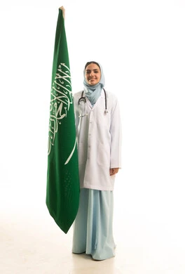 مهن ووظائف طبية، الاحتفال باليوم الوطني السعودي، بورتريه لطبيبة عربية سعودية تلبس رداء وسماعة الطبيب تحمل علم المملكة العربية السعودية، النظر بايماءات الفخر و السعادة ، الجسم بالطول الكامل، خلفية بيضاء