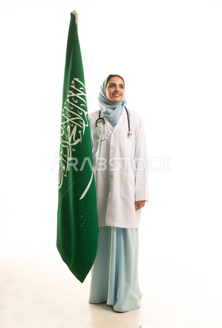 Celebrating Saudi National Day, a portrait of a Saudi Arabian female doctor wearing a coat and stethoscope holding the flag of the Kingdom of Saudi Arabia, looking with gestures of pride and happiness, medical professions and jobs, full-body, white background.