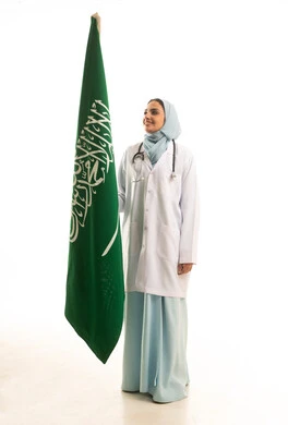 ذكرى توحيد المملكة العربية السعودية، بورتريه لطبيبة عربية سعودية تلبس رداء وسماعة الطبيب تمسك بيديها علم السعودية تحتفل باليوم الوطني بفخر وعزة , العلم السعودي راية التوحيد والاسلام , الجسم بالطول الكامل، خلفية بيضاء