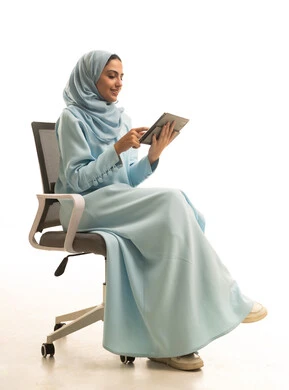 استخدام التقنية الحديثه المتطورة في الحياة العملية عبر الجهاز اللوحي، تعابير وايماءات الاندماج، بورتريه لشابة عربية خليجية سعودية ترتدي الحجاب والعباءة تجلس على كرسي تقوم بالعمل على جهازالتابلت، خلفية بيضاء