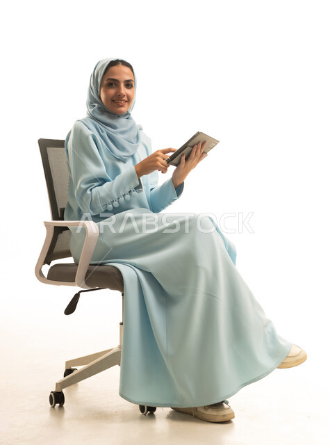 النظر للكاميرا بتعابير السعادة، استخدام التقنية الحديثه المتطورة في الحياة العملية عبر الجهاز اللوحي، بورتريه لشابة عربية خليجية سعودية ترتدي الحجاب والعباءة تجلس على كرسي تقوم بالعمل على جهازالتابلت، خلفية بيضاء