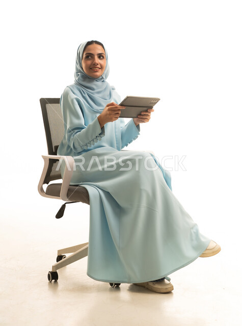 التسوق الالكتروني عبرالأونلاين، استخدام التقنية الحديثه المتطورة في الحياة العملية عبر الجهاز اللوحي، بورتريه لشابة عربية خليجية سعودية ترتدي الحجاب والعباءة تجلس على كرسي تقوم بالعمل على جهازالتابلت، خلفية بيضاء
