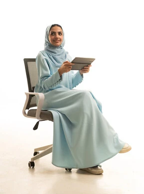 التسوق الالكتروني عبرالأونلاين، استخدام التقنية الحديثه المتطورة في الحياة العملية عبر الجهاز اللوحي، بورتريه لشابة عربية خليجية سعودية ترتدي الحجاب والعباءة تجلس على كرسي تقوم بالعمل على جهازالتابلت، خلفية بيضاء