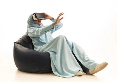 Integration, interaction, and simulation of the imaginary world, a portrait of a Saudi Arabian Gulf woman wearing a hijab and an abaya sitting on a chair between bags, wearing 3D glasses, raising her hand upwards with expressions of integration and stopping an action, white background.