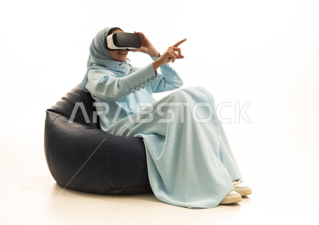 المتعة بالمشاهدة عبر نظارة الواقع الافتراضي، الاشارة الى مكان معين, بورترية لإمراة عربية خليجية سعودية محجبة ترتدي عباءة تجلس على كرسي بين باج تضع نظارات الفي آر, محاكاة العالم الخيالي, الاشارة باصبع السبابة جانبا بتعابير هذا المكان هنا، خلفية بيضاء