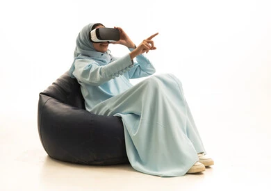 The enjoyment of watching through virtual reality glasses, pointing to a specific place, a portrait of a Saudi Arabian Gulf woman wearing a hijab and an abaya sitting on a chair between bags with VR glasses, simulating the imaginary world, pointing with the index finger to the side with expressions of this place here, white background.