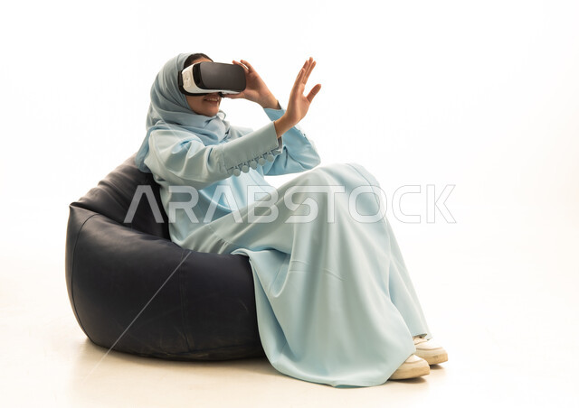 The enjoyment and entertainment of watching through virtual reality glasses, a portrait of a Saudi Arabian Gulf woman wearing a hijab and an abaya sitting on a chair between bags, wearing 3D glasses, raising her hand upwards with expressions of engagement and stopping from doing something, white background.